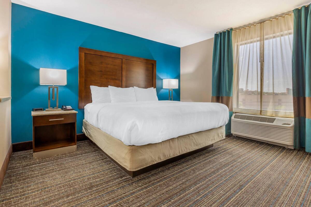A hotel room with a large bed, two bedside tables with lamps, a window with curtains, and a wall heater.