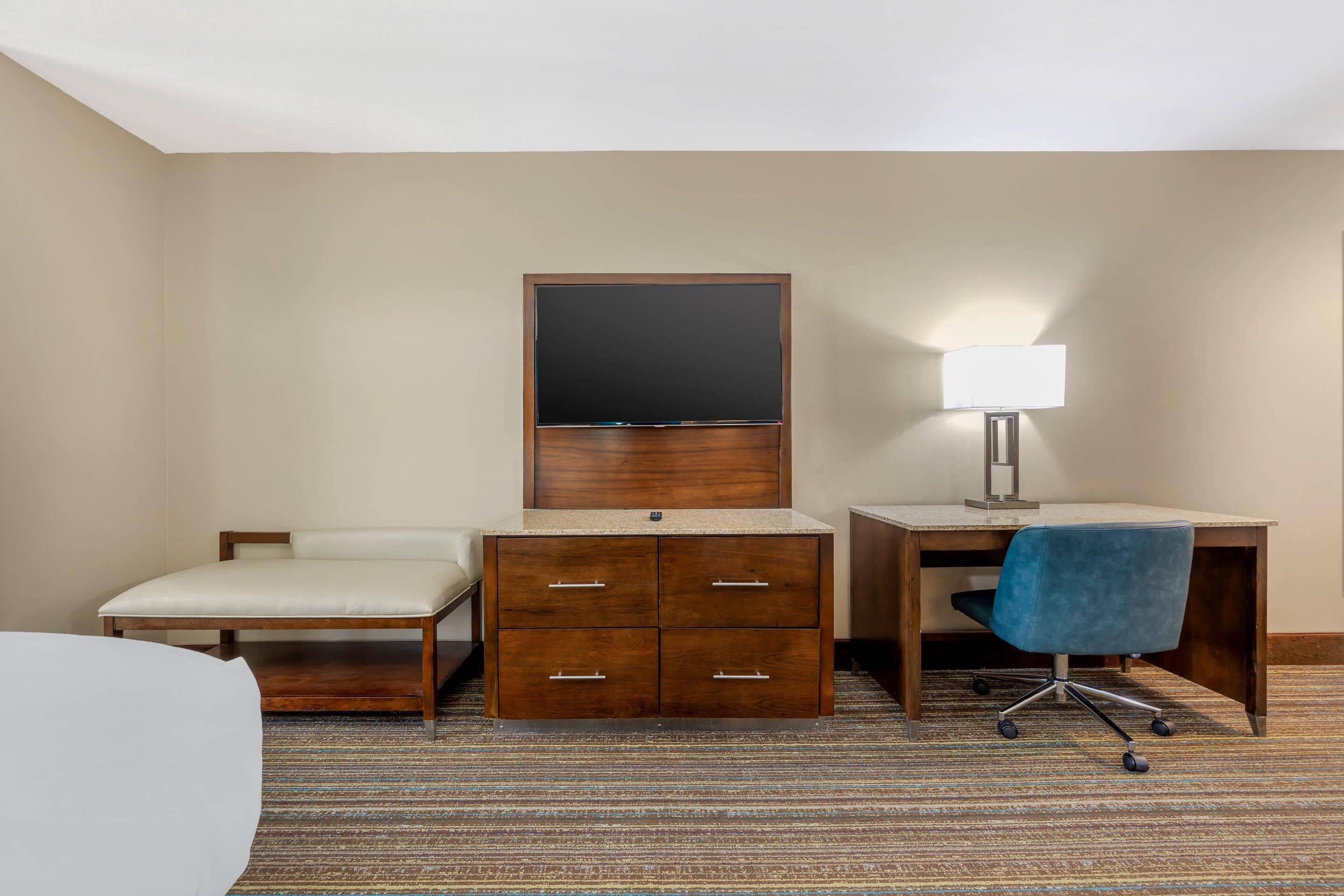 A hotel room with a bed, a desk with a lamp, a chair, a wardrobe, and a television.