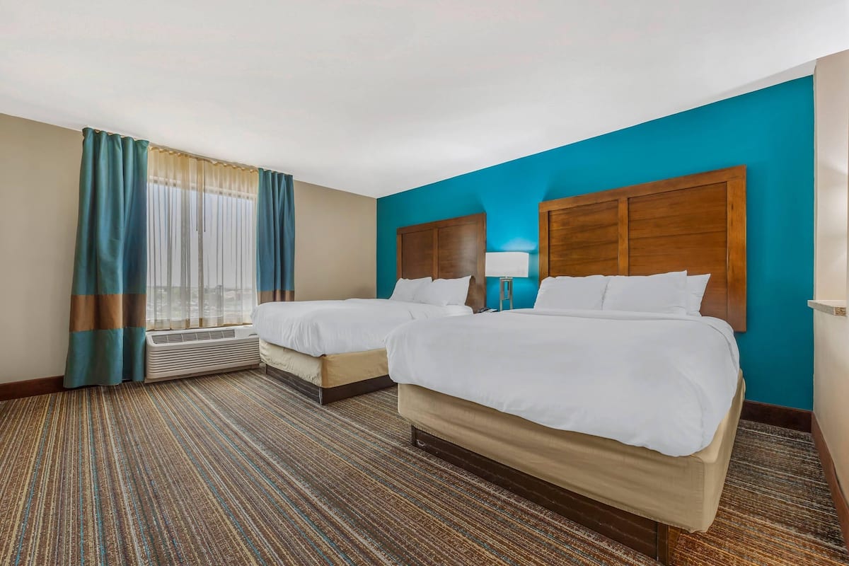 A hotel room with two beds, a large window with curtains, a carpeted floor, and a blue accent wall.