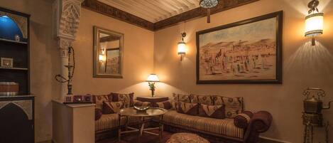 A traditional room with a sofa, a round coffee table, a patterned ottoman, and a painting on the wall.