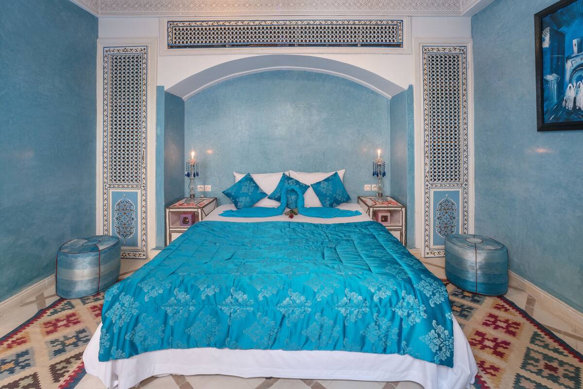 A large bed with a blue bedspread, two bedside tables with lamps, and a patterned rug on the floor.