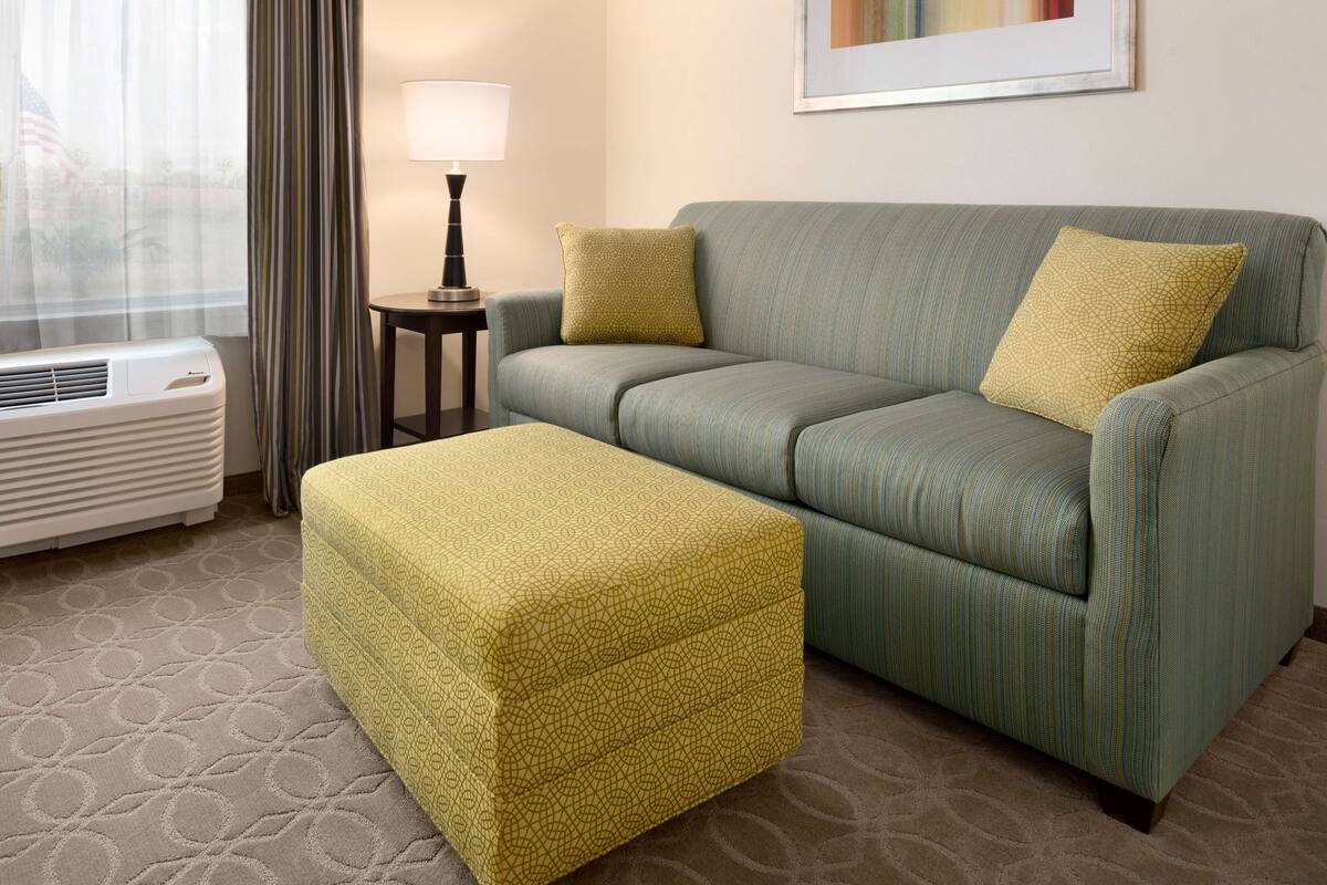 A hotel room with a green sofa, a matching ottoman, a lamp, and a framed picture on the wall.
