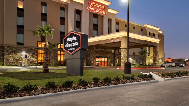 Edifício do Hampton Inn & Suites by Hilton com fachada de pedra, entrada bem iluminada e jardim frontal ajardinado.