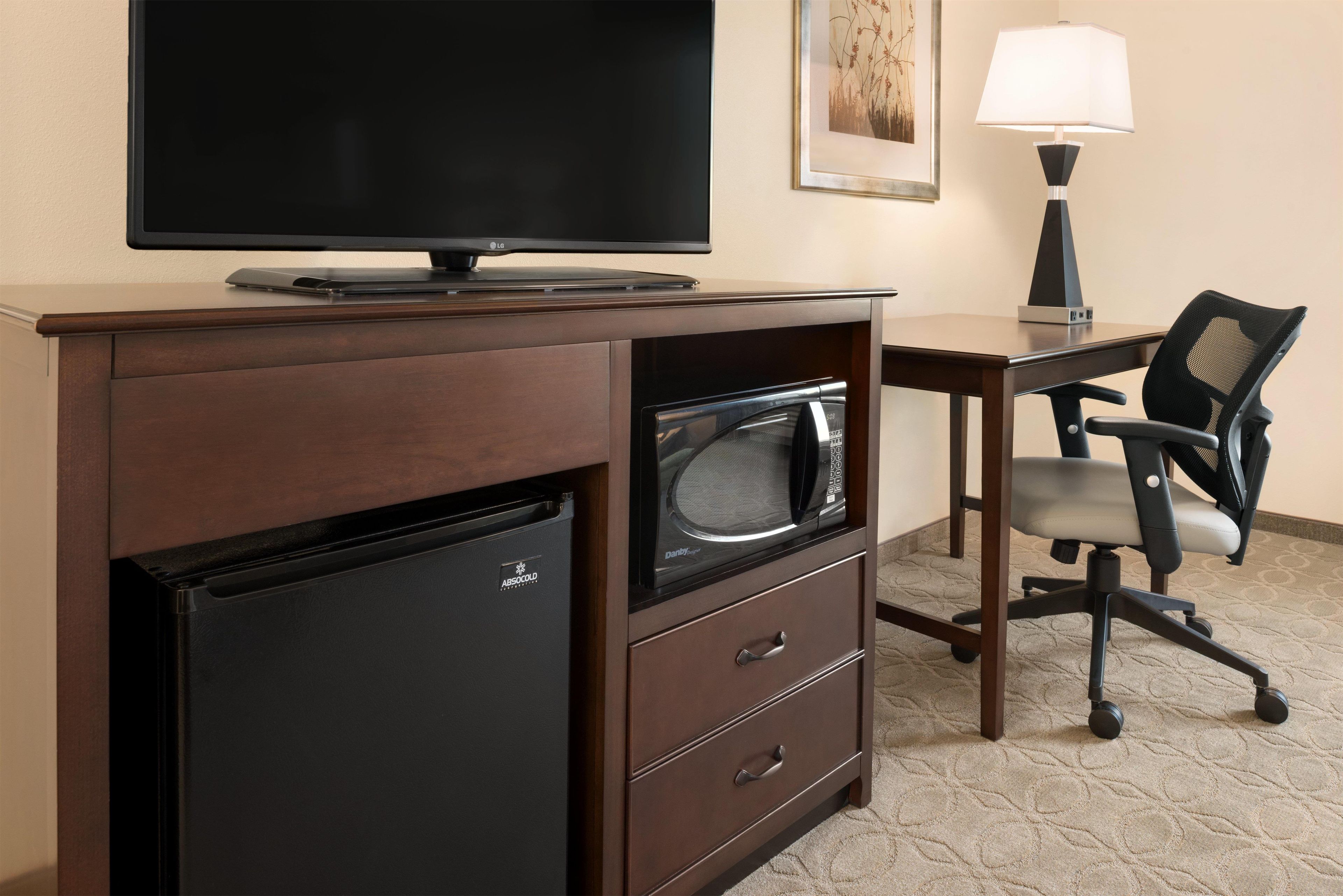 A hotel room with a flat-screen TV, a microwave, a mini-fridge, a desk, a chair, and a lamp.