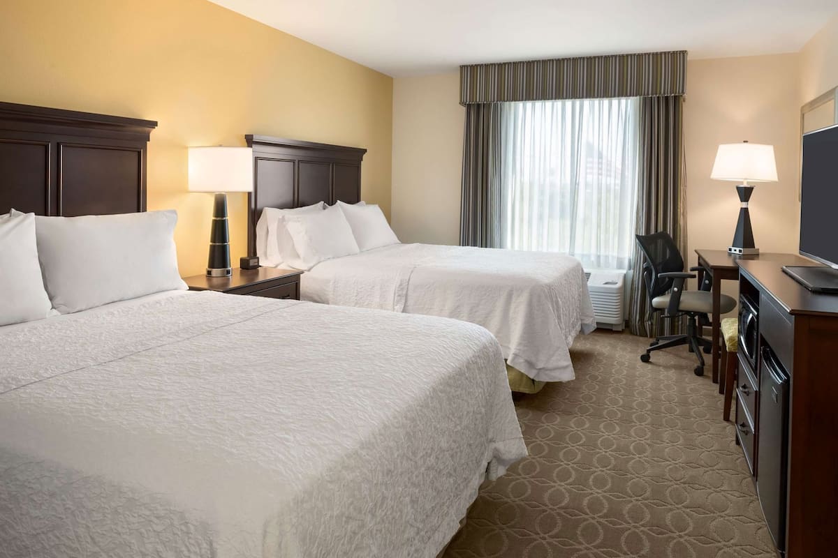 A hotel room with two beds, a desk, a chair, a TV, and a window with curtains.
