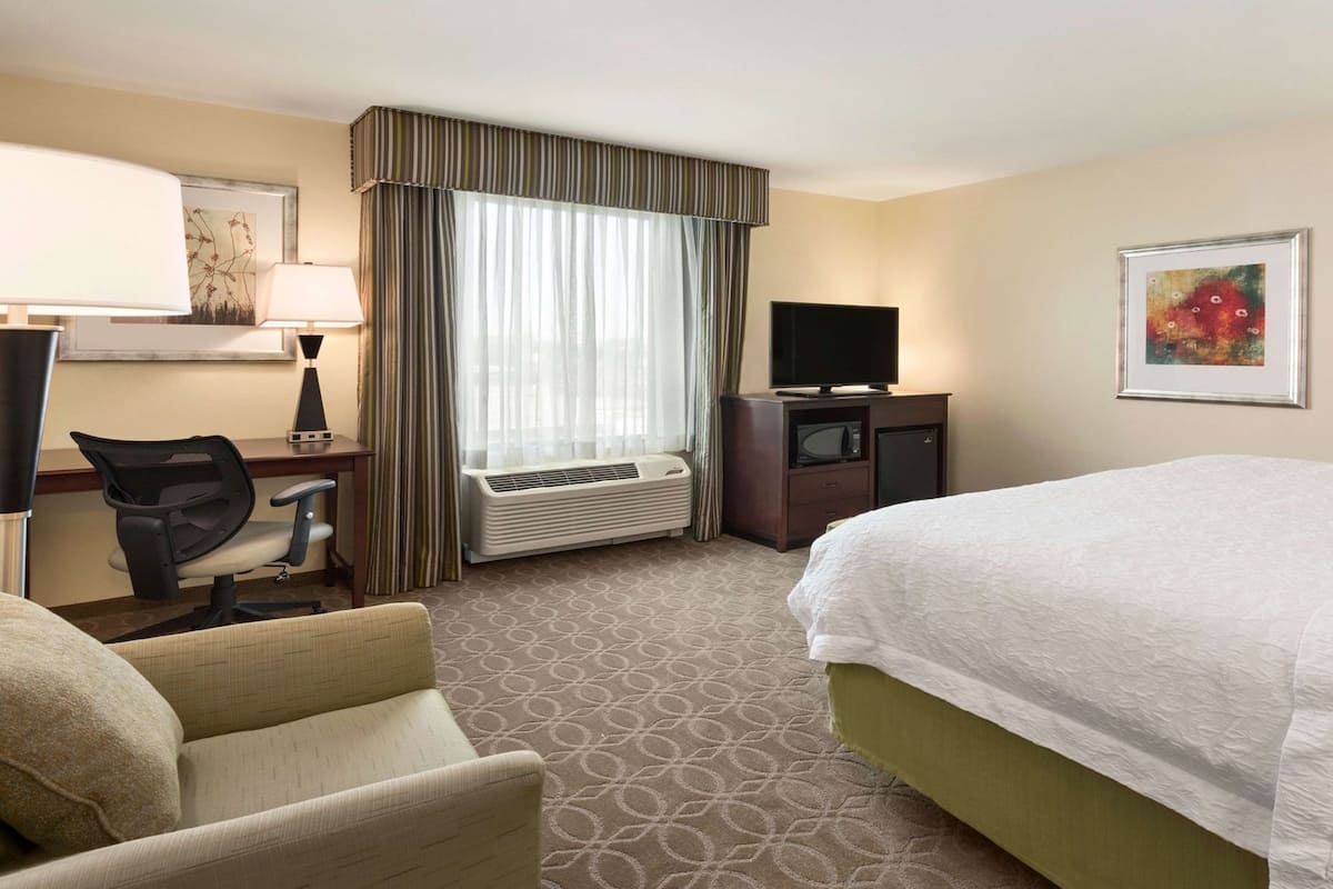 A hotel room with a bed, a desk with a chair, a television, an air conditioning unit, and a window with curtains.