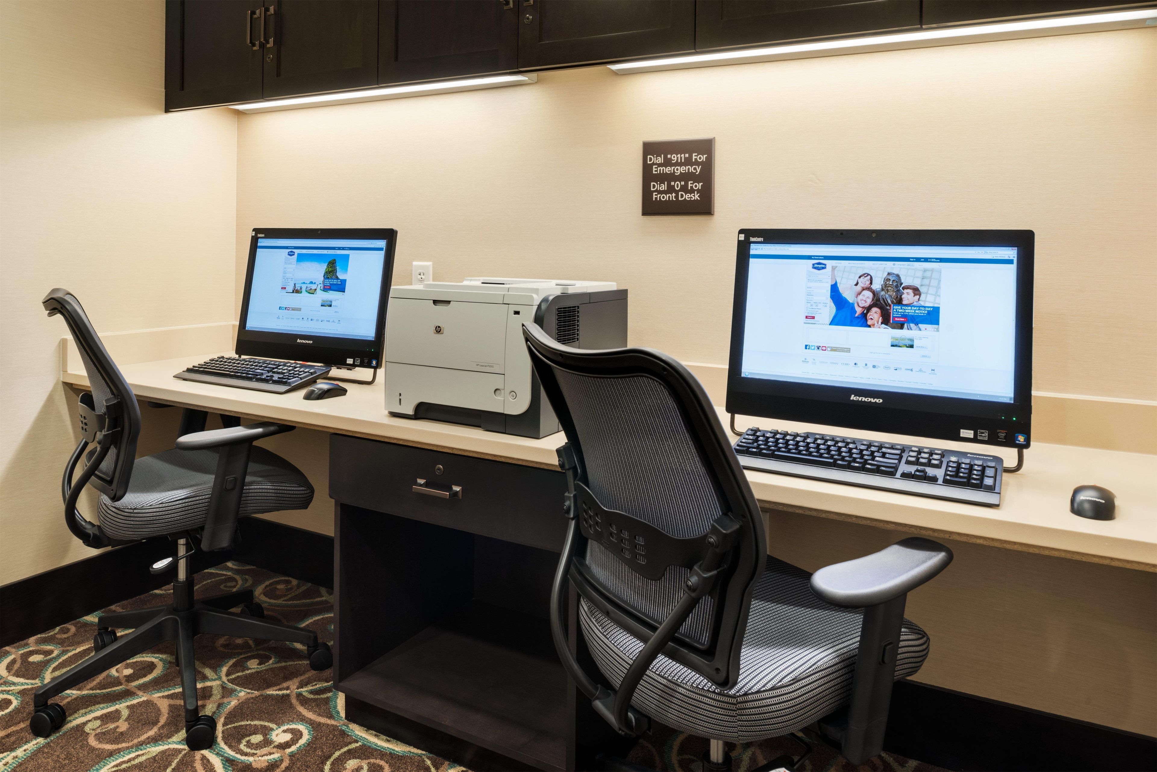 A hotel workspace with two Lenovo computers, a printer, and a sign indicating emergency and front desk contact numbers.