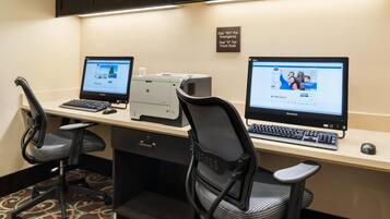 A hotel workspace with two Lenovo computers, a printer, and a sign indicating emergency and front desk contact numbers.