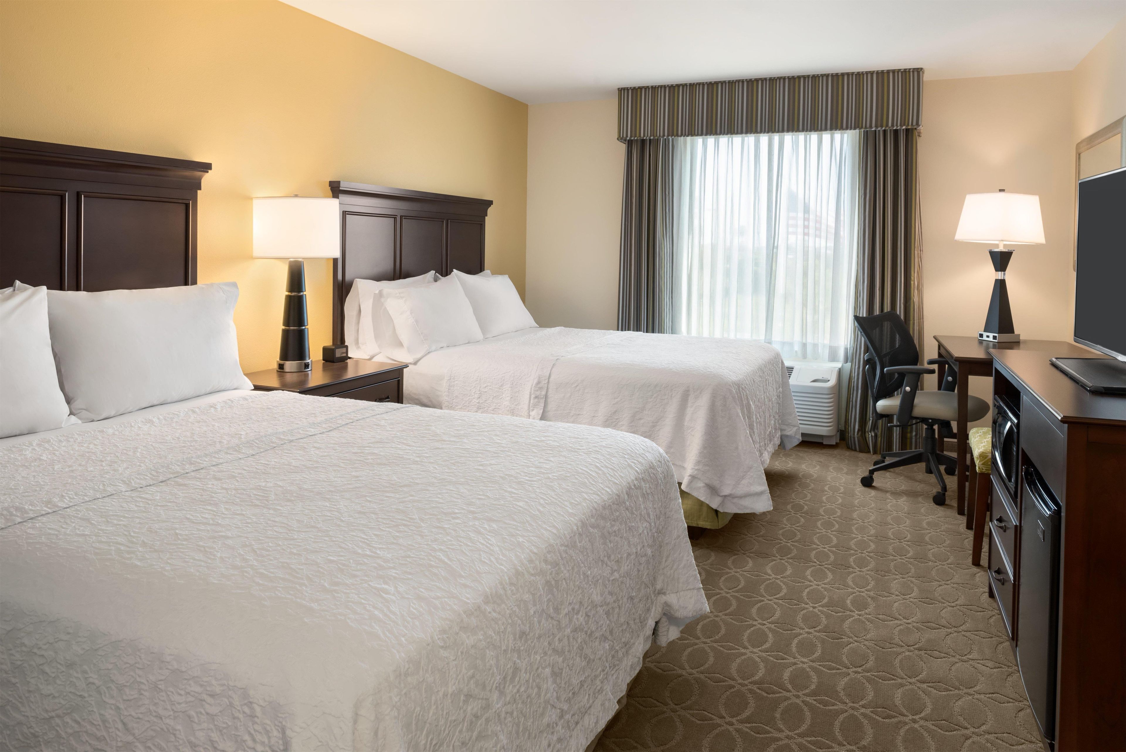 A hotel room with two beds, a desk, a chair, a television, and a window with curtains.