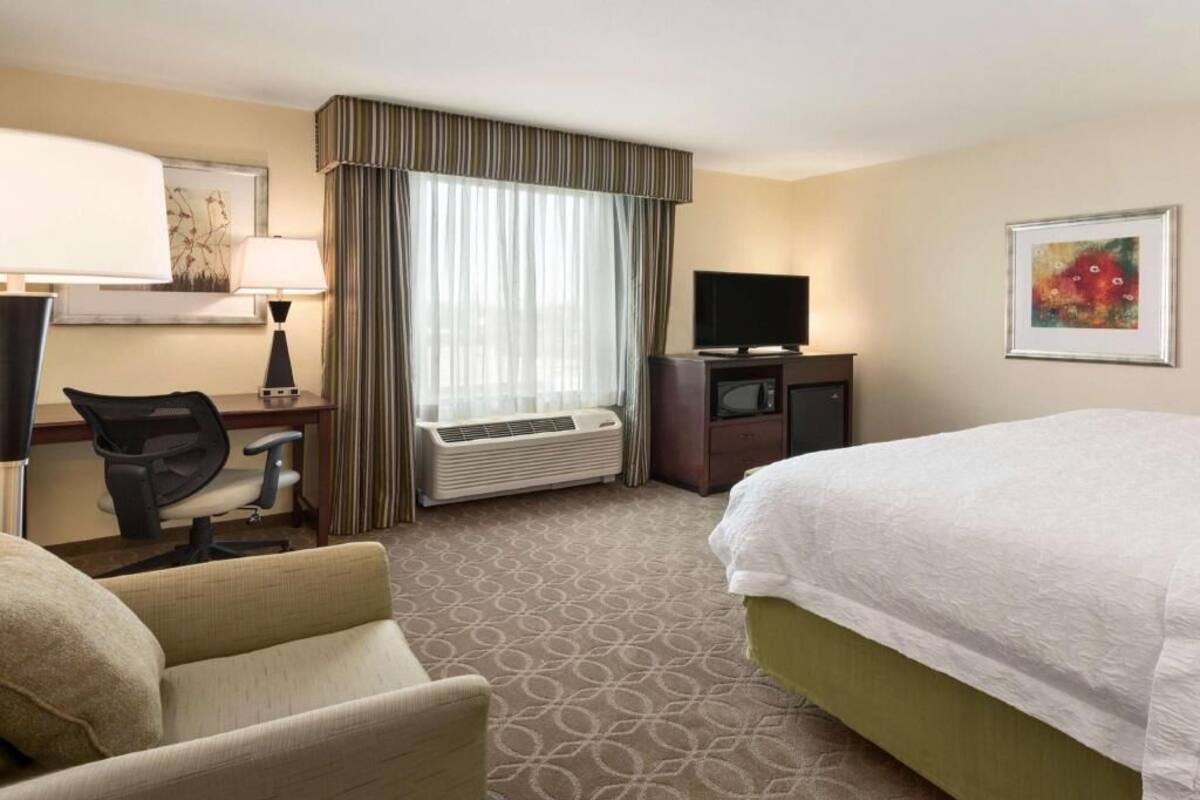 A hotel room with a bed, a desk with a chair, a television, an air conditioning unit, and a window with curtains.