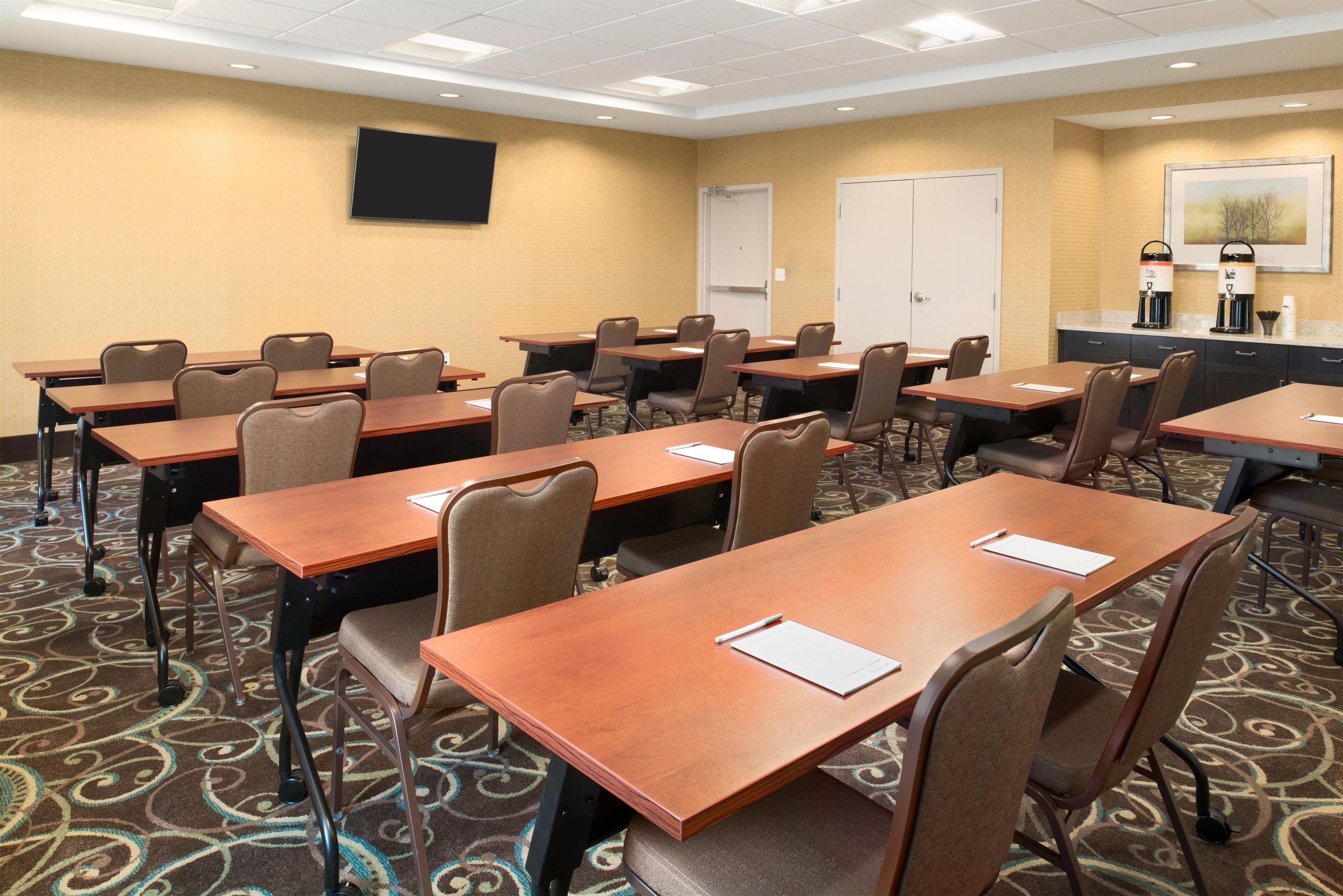 A conference room with multiple tables and chairs, a flat-screen TV mounted on the wall, and a coffee station with two coffee makers.
