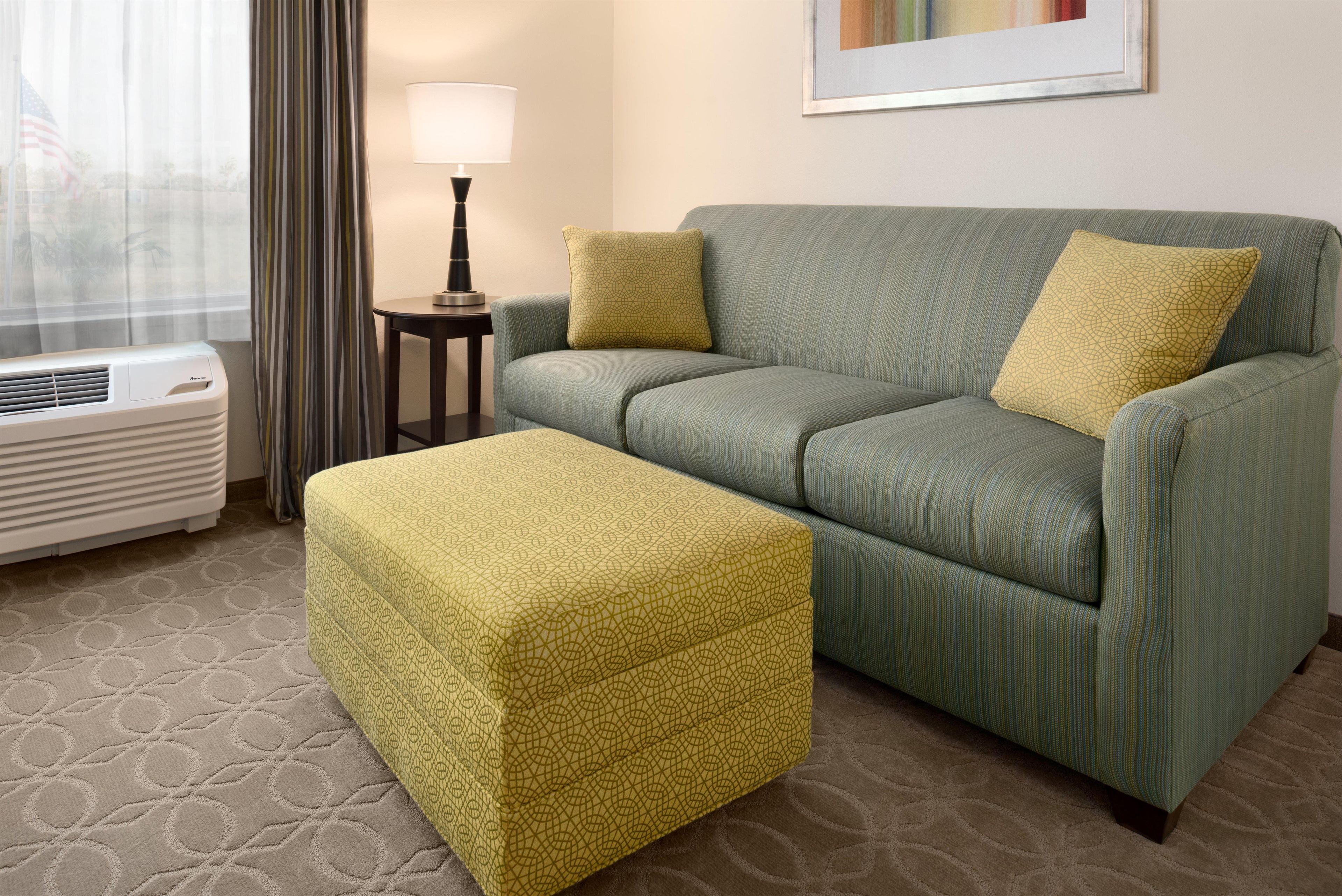 Ein Hotelzimmer mit einem grünen Sofa, einem passenden Hocker, einer Lampe auf einem Beistelltisch und einem Fenster mit Vorhängen.