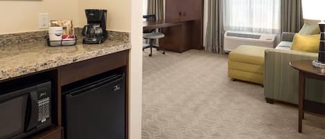 Habitación de hotel compacta con kitchenette, zona de estar con sofá y sillón, y un gran ventanal con cortinas.