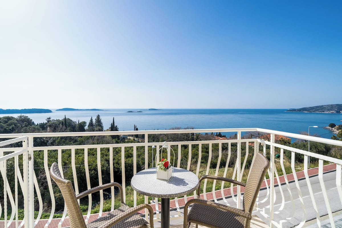A balcony with wicker chairs, a small table with a vase of flowers, and a view of the sea and distant land.