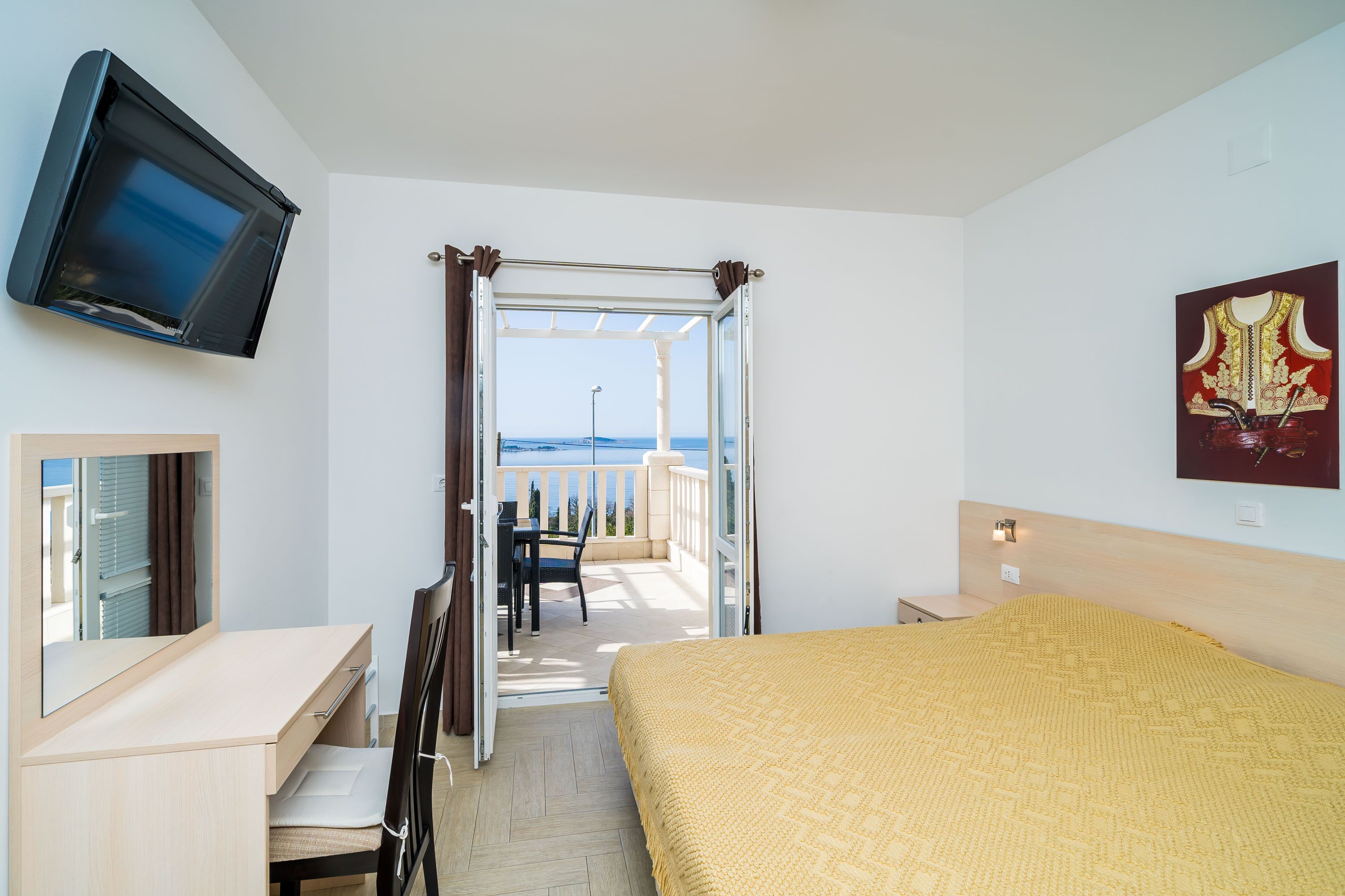 Une chambre d’hôtel avec un lit, un bureau, une télévision, un balcon donnant sur la mer et un tableau au mur.