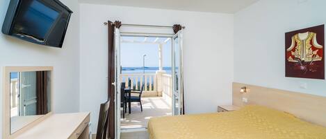 Une chambre d’hôtel avec un lit, un bureau, une télévision, un balcon donnant sur la mer et un tableau au mur.