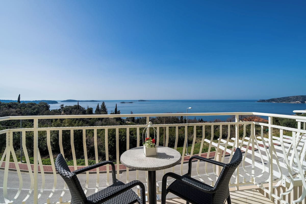 A balcony with wicker chairs and a small table, overlooking a scenic view of the sea and hills.