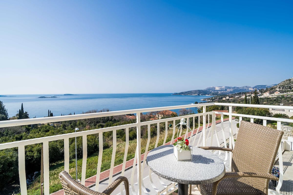 A balcony with wicker furniture, a table with a vase of flowers, and a view of the sea and distant hills.
