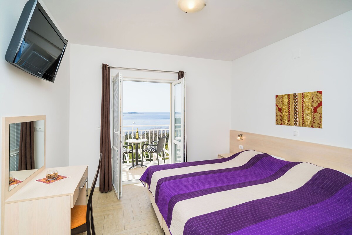 A hotel room with a bed, a desk, a chair, a TV, and a balcony with a table and chairs.