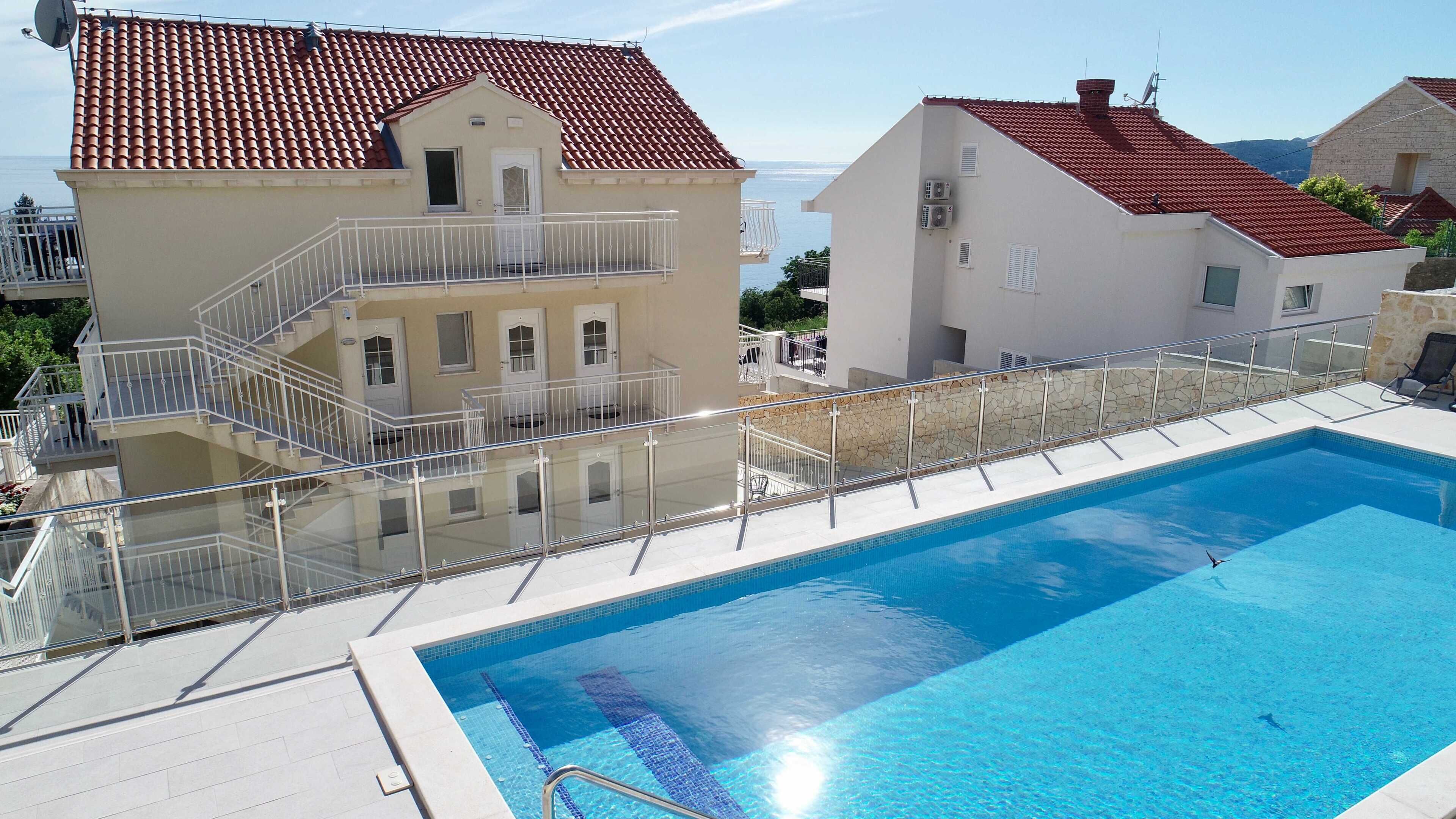 Una piscina con barandilla de vidrio, un edificio con techo de tejas rojas y fachada blanca.
