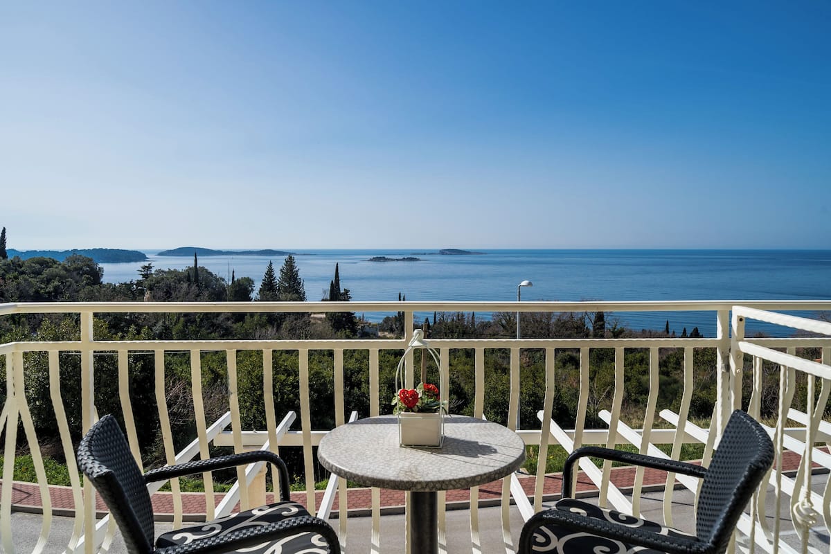 A balcony with a round table and two chairs, overlooking a scenic view of the sea and distant islands.