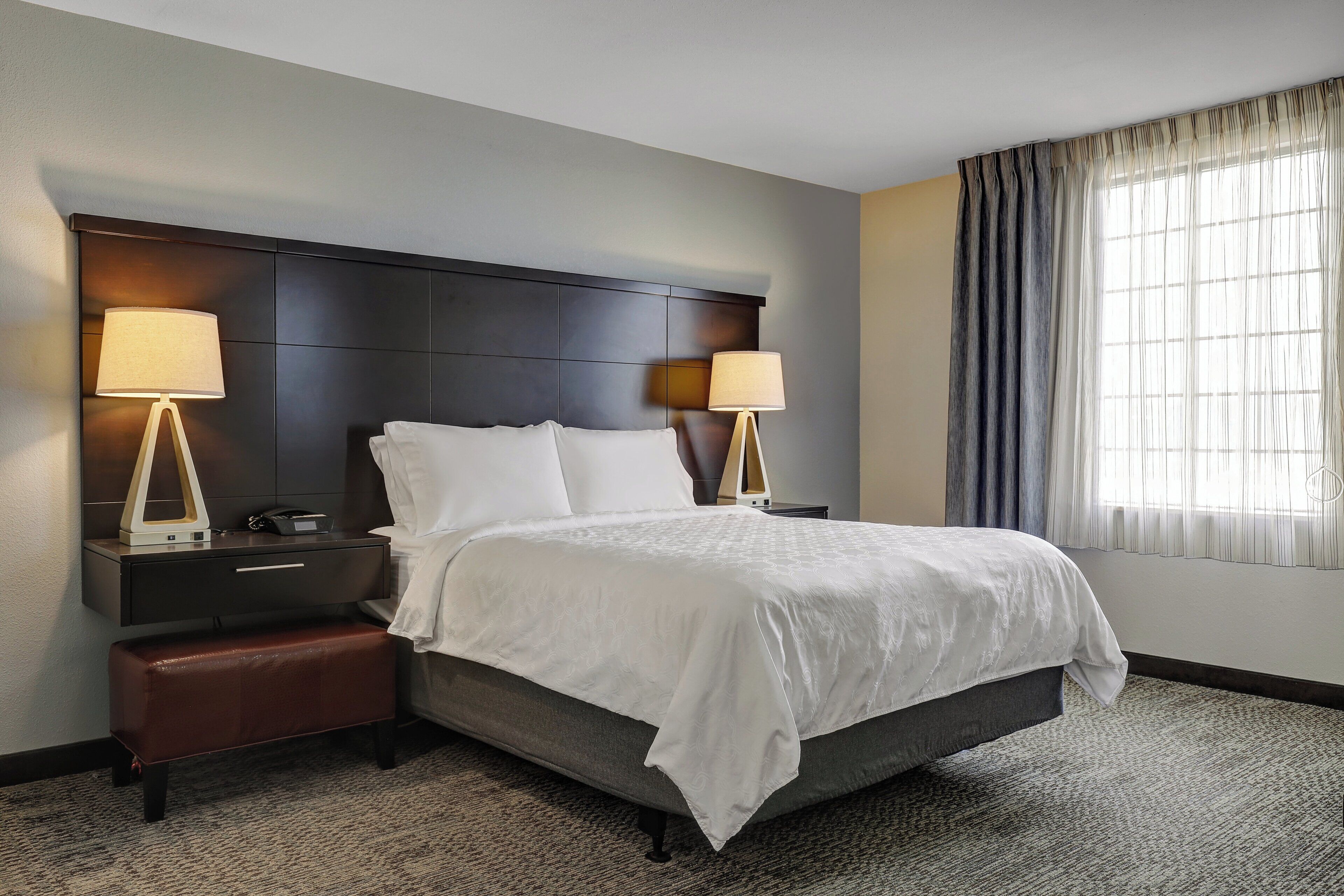 A hotel room with a large bed, two bedside tables with lamps, a bench, a headboard, and a window with curtains.