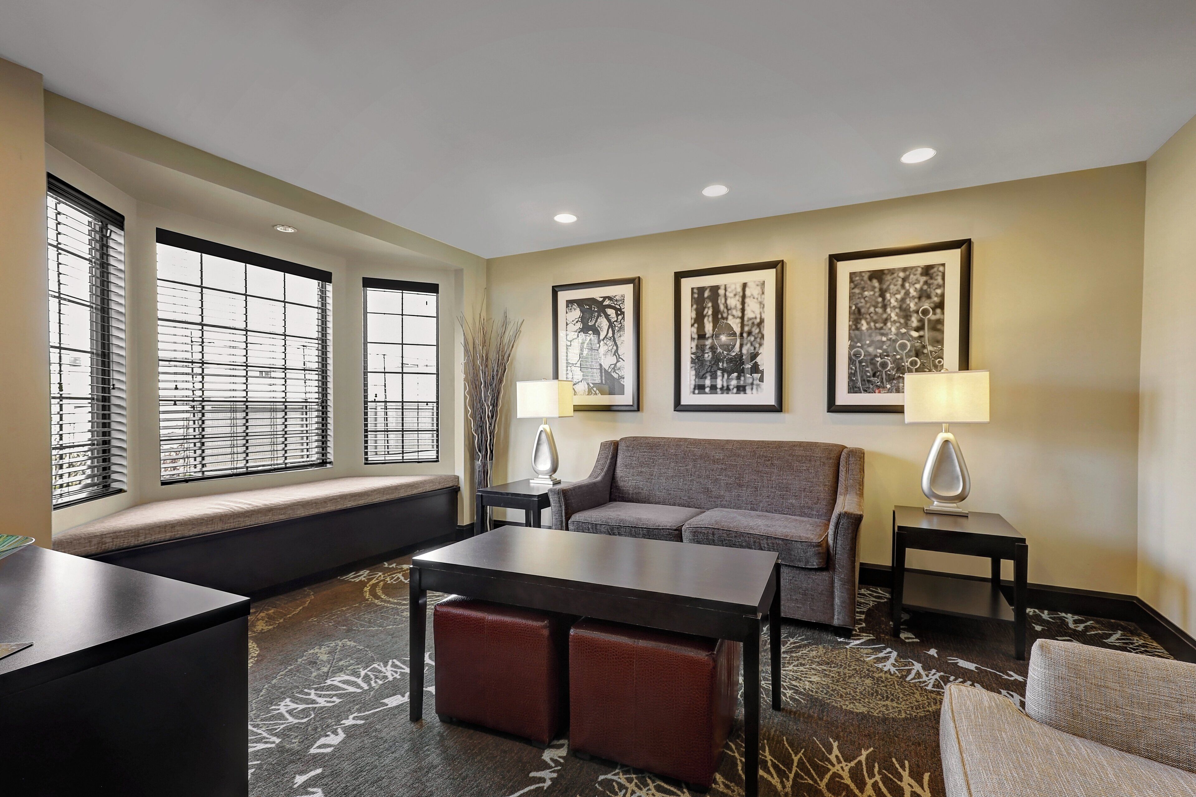 A hotel room with a sofa, a small table, a bench, and a window with blinds. There are two lamps on side tables and three framed pictures on the wall.