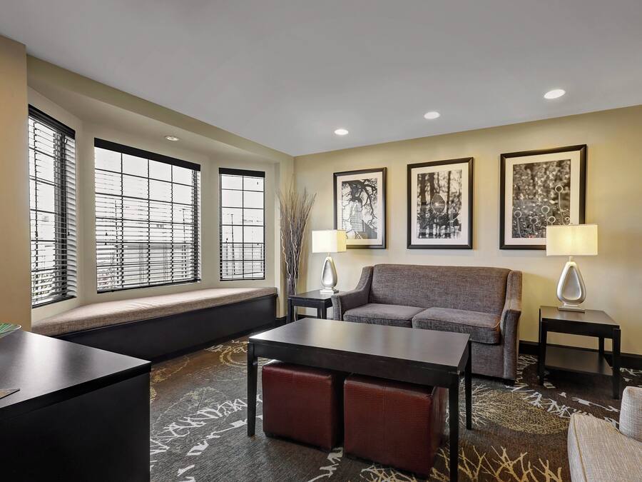 A hotel room with a sofa, a small table, a bench, and a window with blinds. There are two lamps on side tables and three framed pictures on the wall.