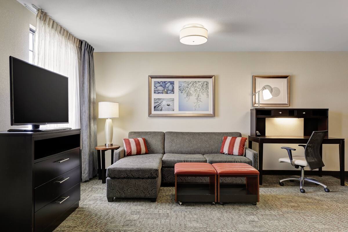 A hotel room with a flat-screen TV, a sofa, a desk with a chair, a lamp, and framed artwork on the wall.