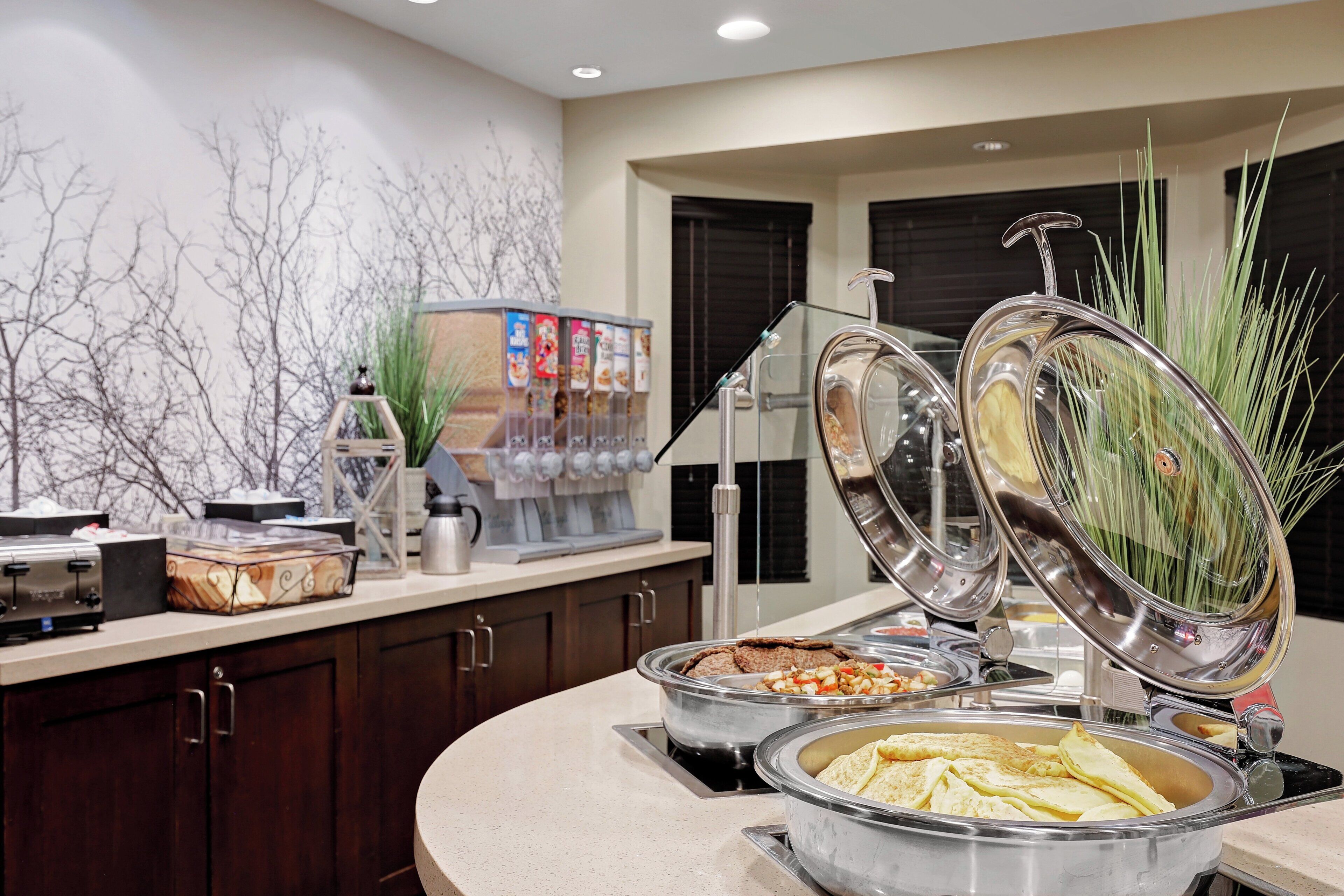 A buffet setup with various food items, a coffee machine, and a decorative wall with a tree design.
