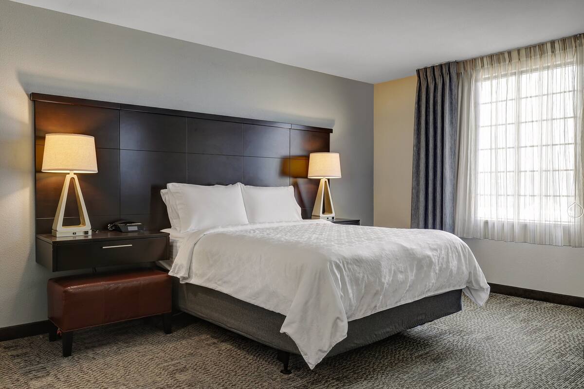 A hotel room with a large bed, two bedside tables, a bench, and a window with curtains.
