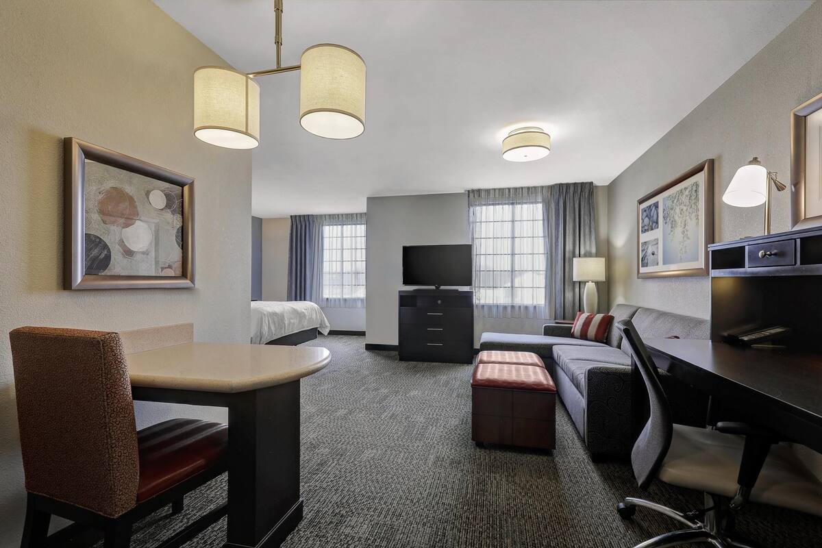 A hotel room with a bed, a desk, a chair, a sofa, a TV, and artwork on the walls.