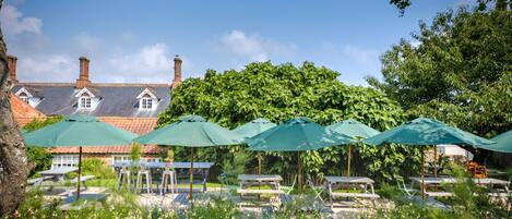 A garden with picnic tables and umbrellas, a brick building with chimneys, and lush greenery in the background.