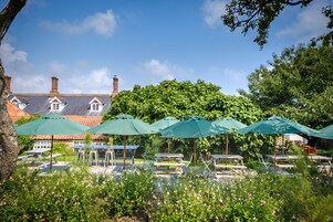 A garden with picnic tables and umbrellas, a brick building with chimneys, and lush greenery in the background.