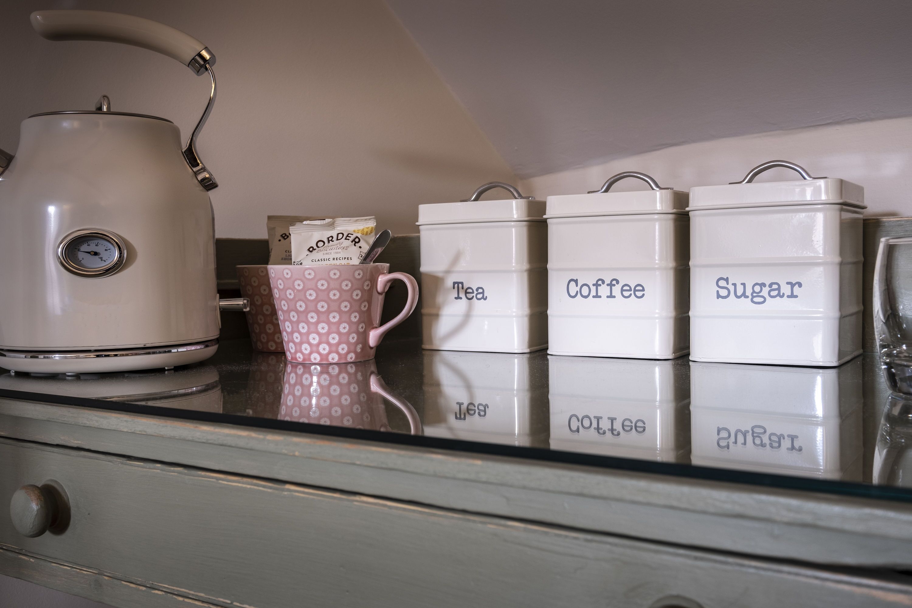 A kitchen counter with a kettle, a polka-dotted mug, and labeled containers for tea, coffee, and sugar.