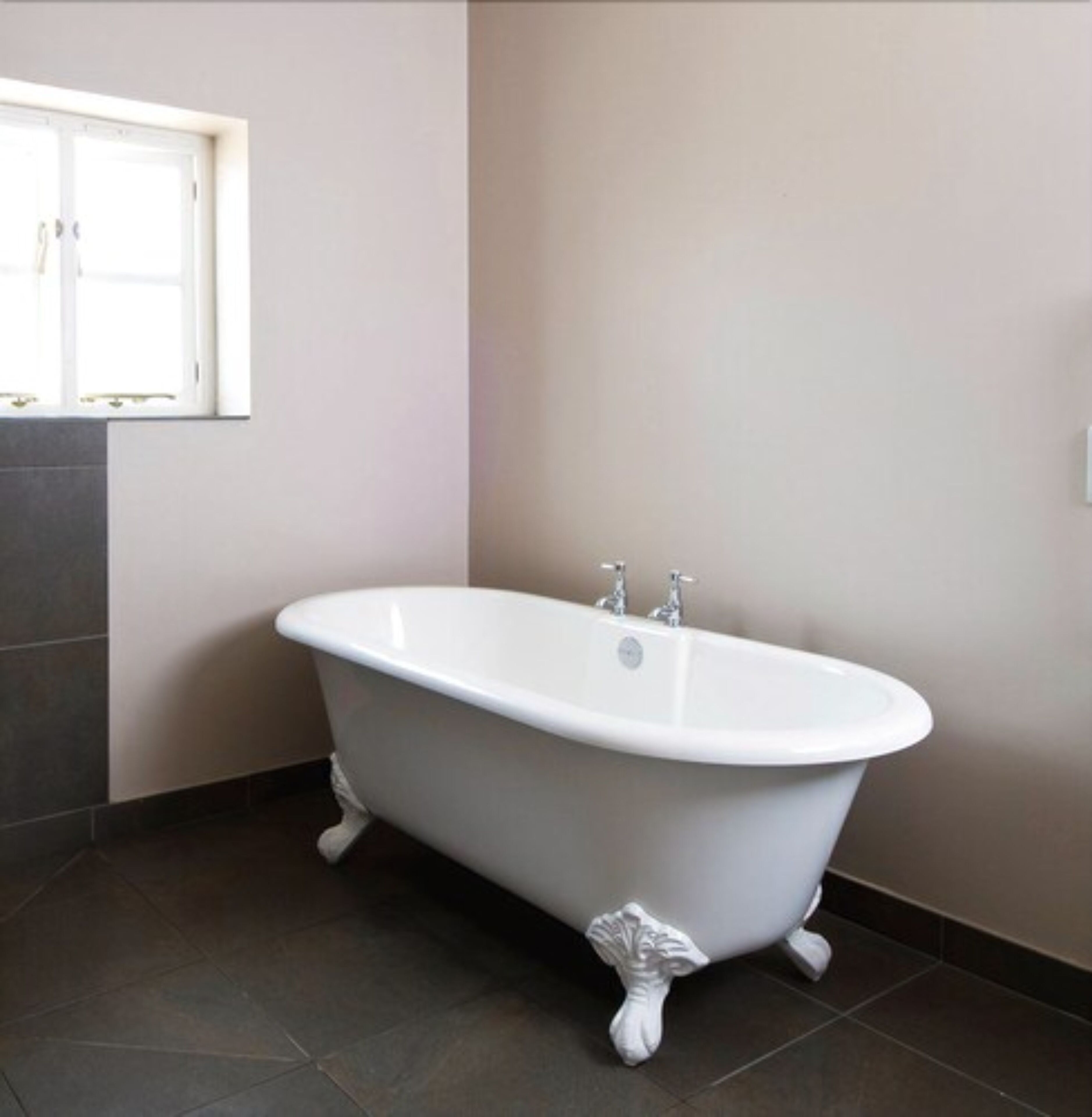 Eine freistehende Badewanne in einem Badezimmer mit dunkel gefliestem Boden und einem Fenster darüber.