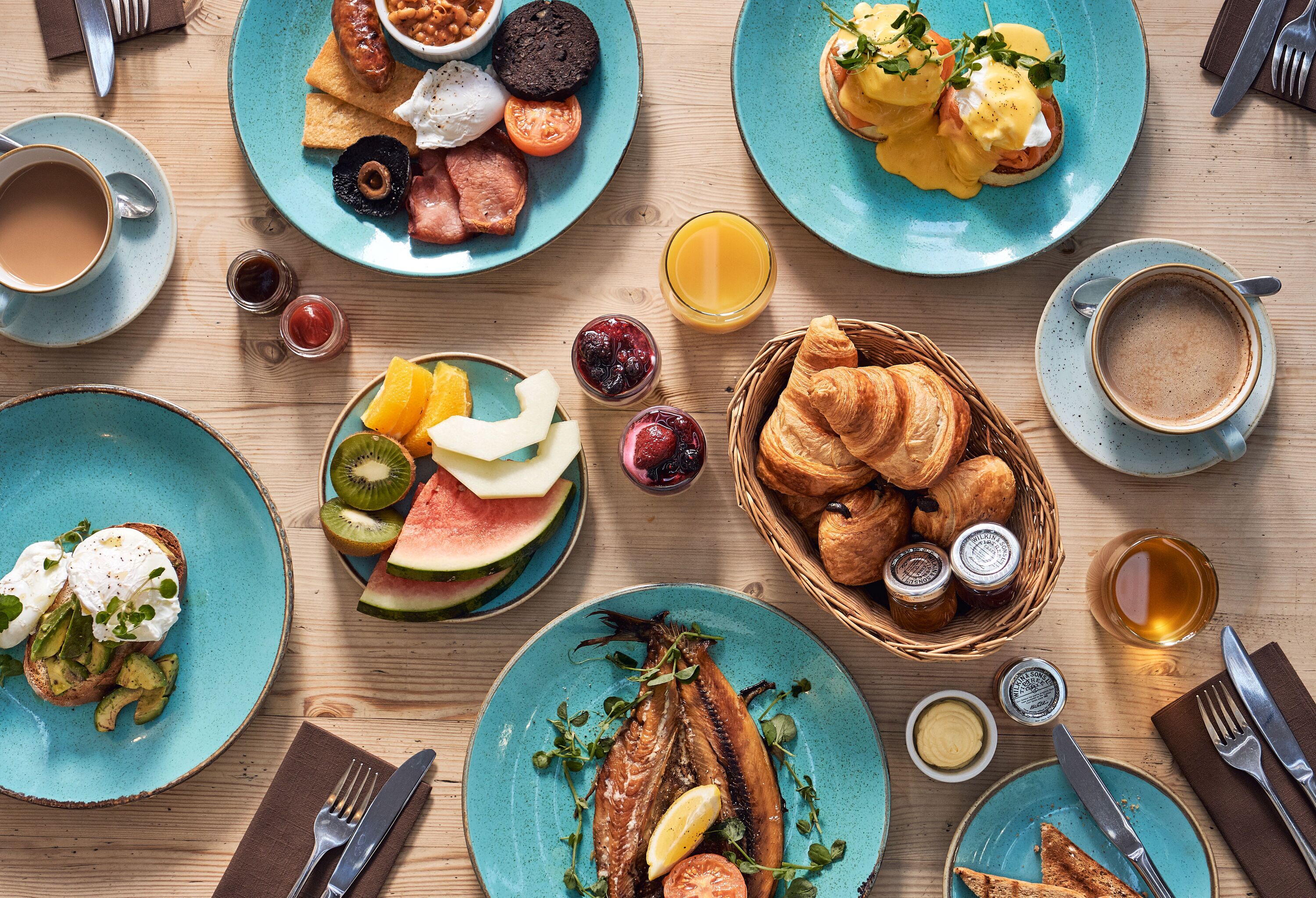 A breakfast setting with various dishes, including eggs, toast, fruits, and pastries, arranged on a wooden table.