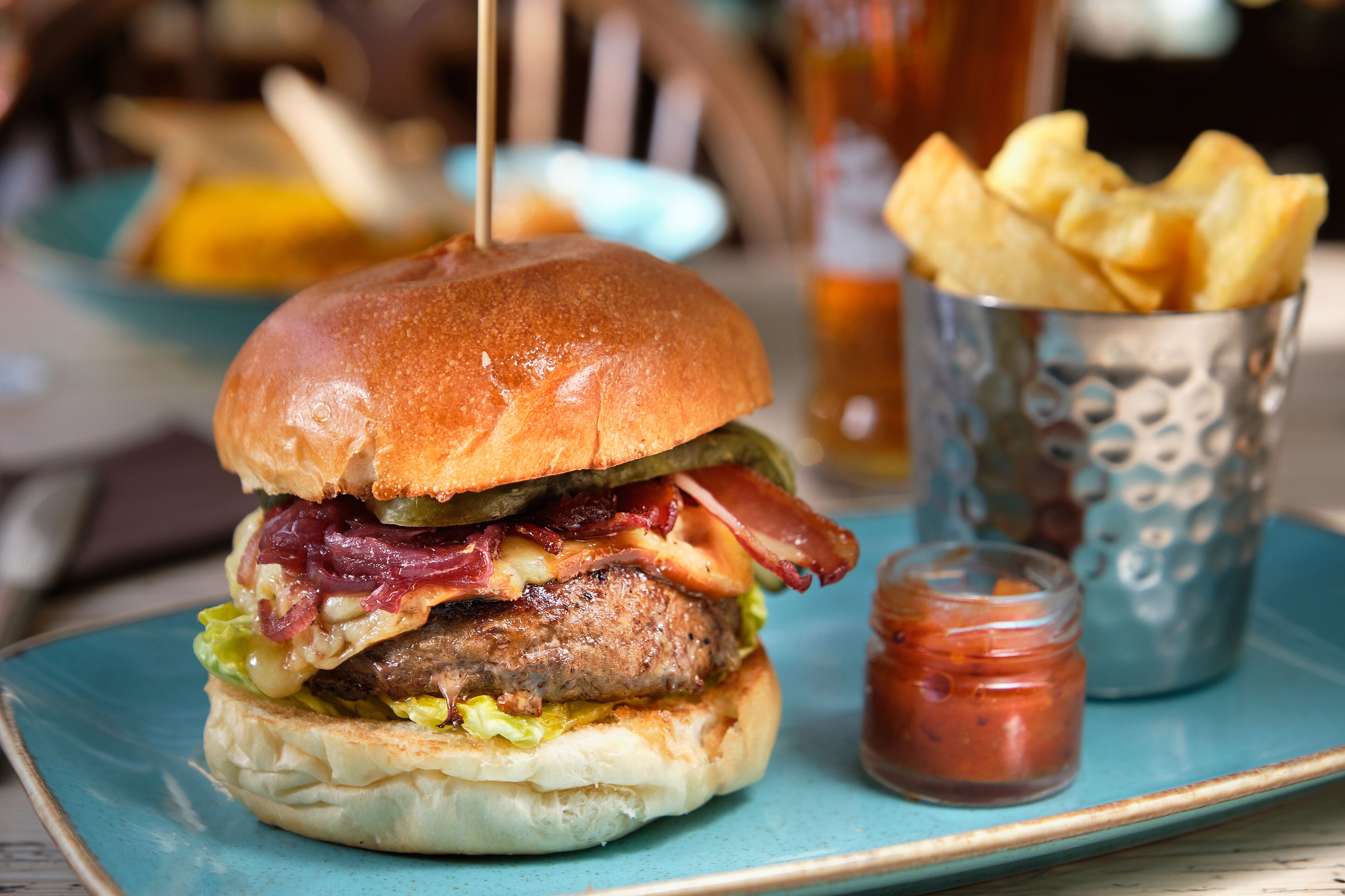 A burger with a bun, bacon, cheese, and lettuce, served on a blue plate with a side of fries and a small jar of ketchup.