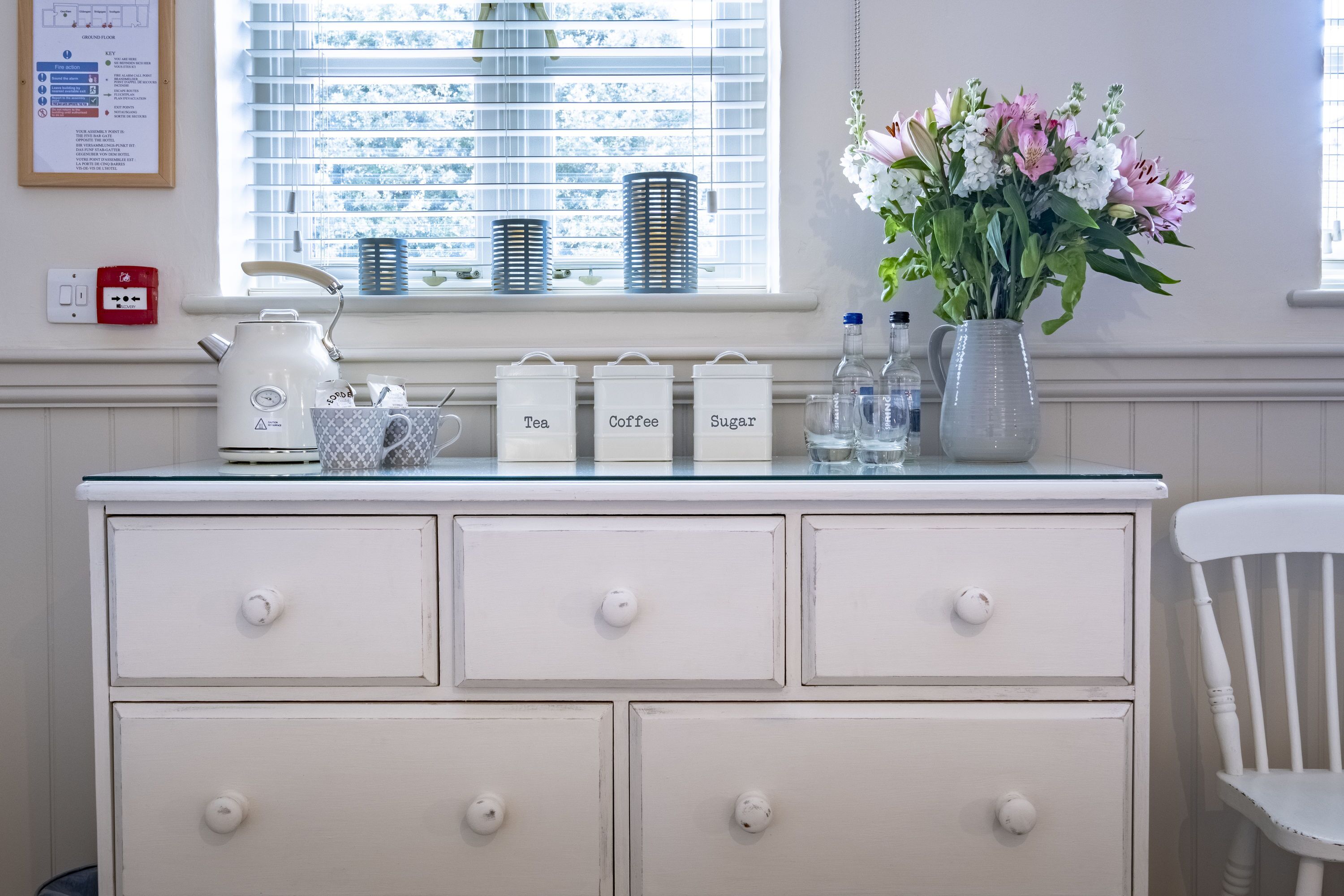 Ein weißer Kommode mit beschrifteten Behältern für Tee, Kaffee und Zucker, einem Wasserkocher und einer Vase mit Blumen.