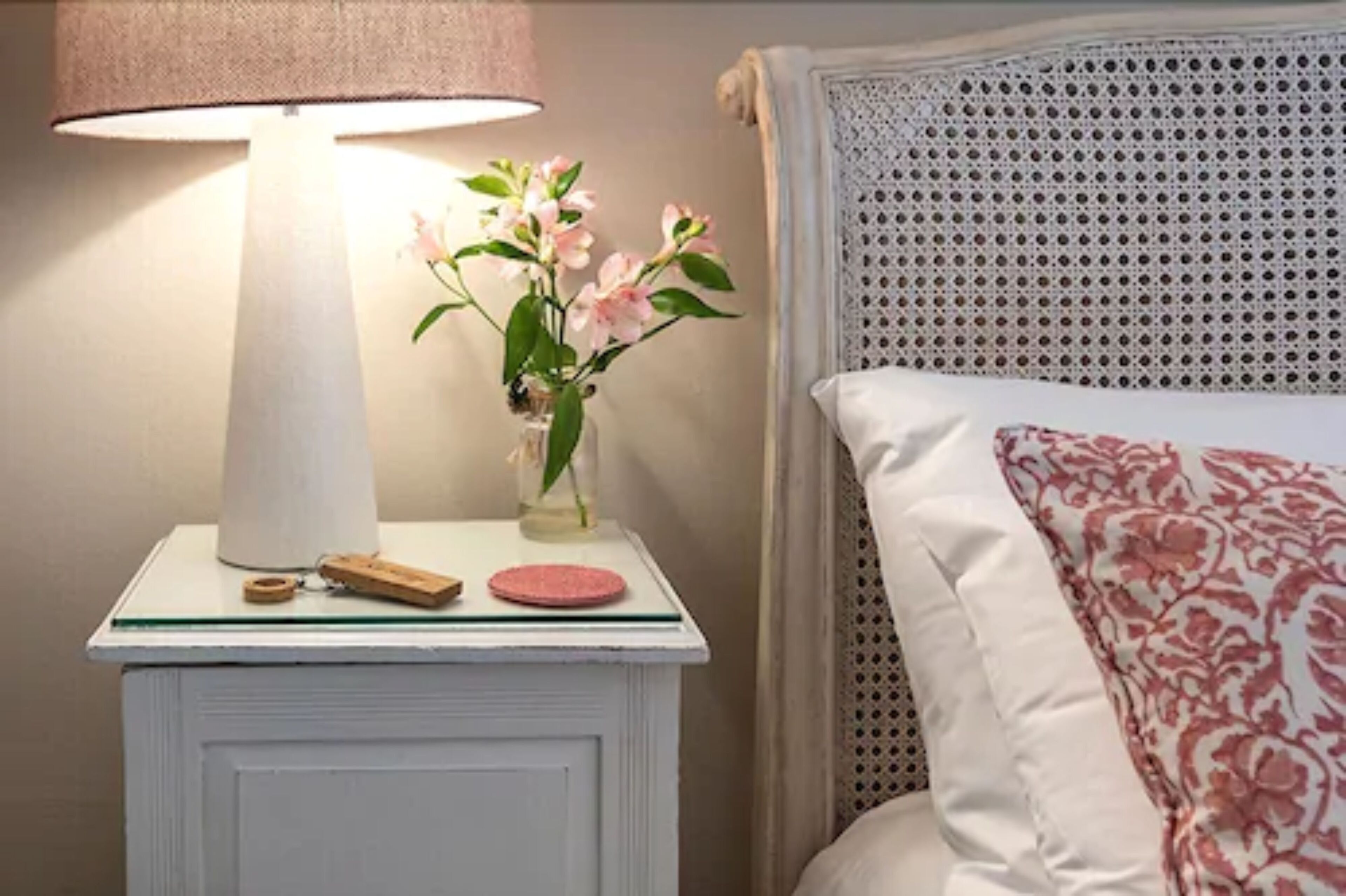 A neatly made bed with a patterned pillow and a bedside table with a lamp, a vase with flowers, and a small wooden object.