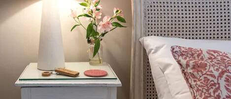A neatly made bed with a patterned pillow and a bedside table with a lamp, a vase with flowers, and a small wooden object.