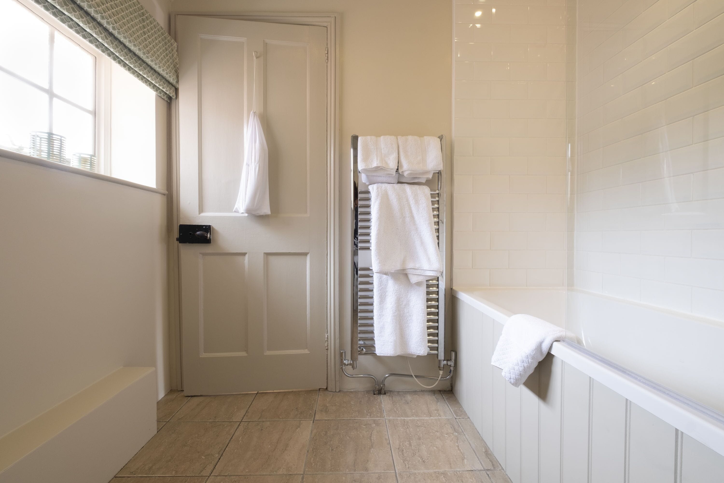 Un baño con una puerta blanca, un perchero con varias toallas, una bañera y una ventana con persianas.