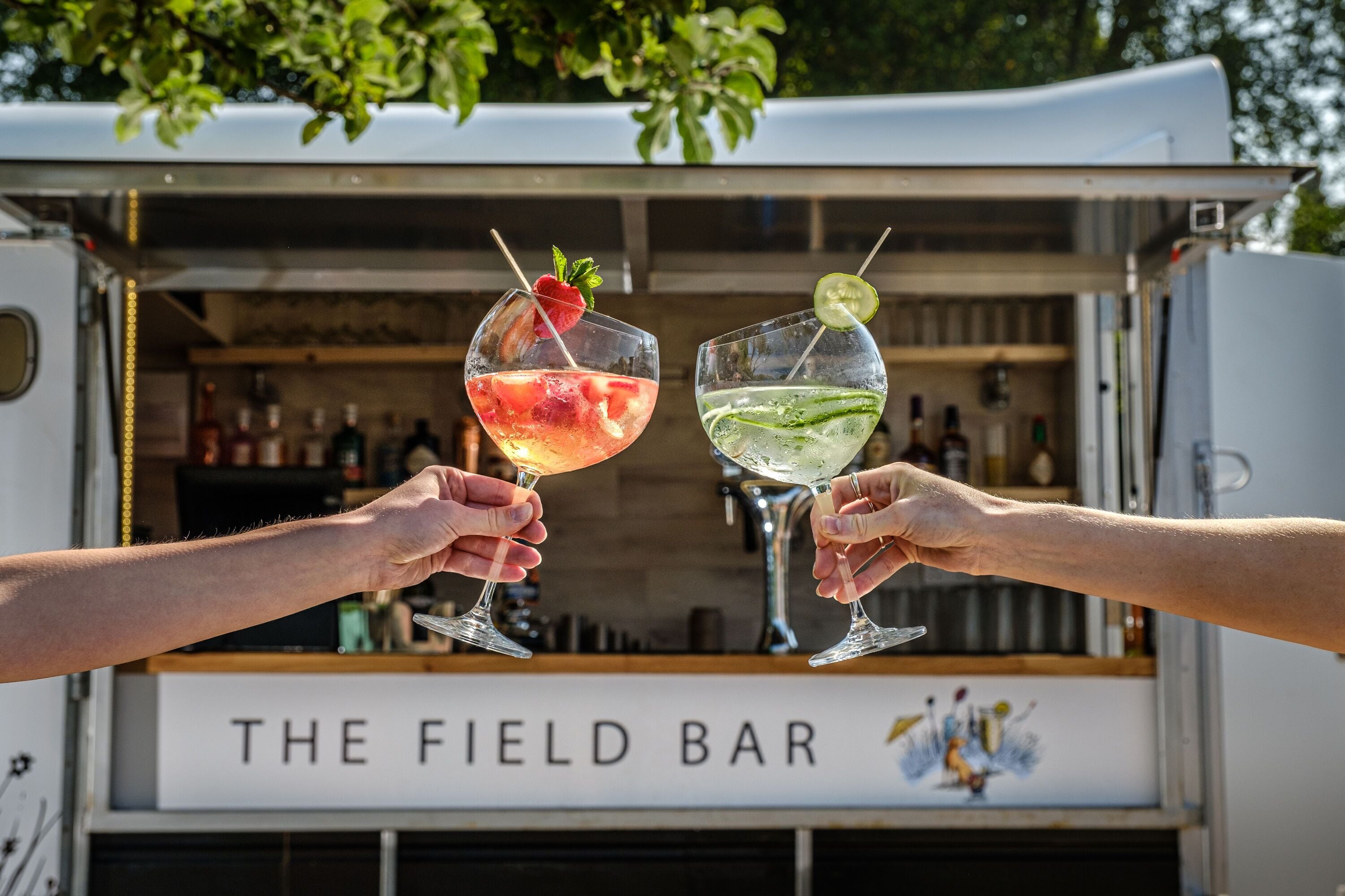 Two people holding drinks in front of a food truck labeled "THE FIELD BAR".