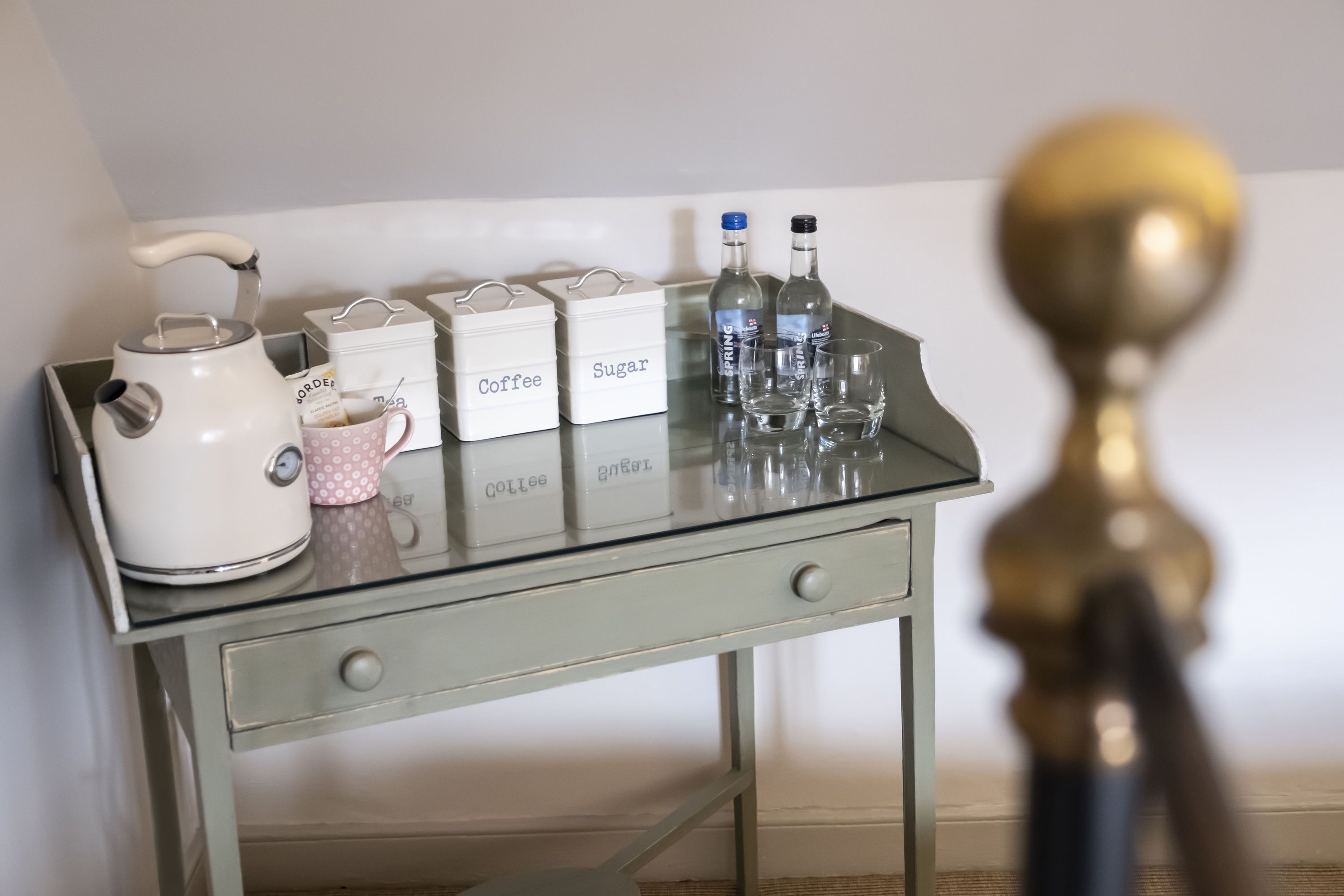 A glass-top table with labeled containers for coffee, sugar, and tea, a kettle, and a polka-dotted mug.