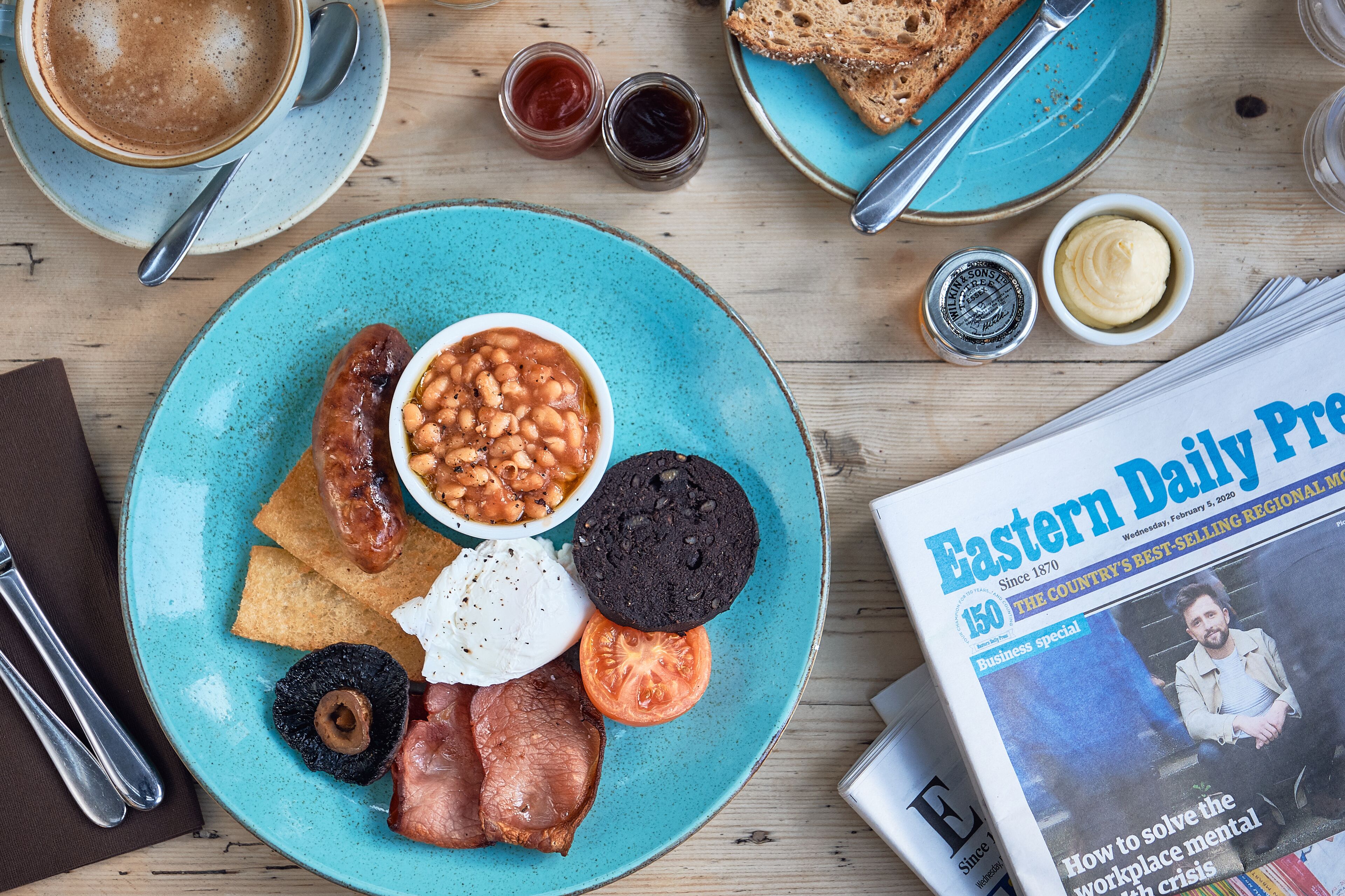 A breakfast plate with beans, sausage, bacon, eggs, and toast, along with a newspaper and coffee on a wooden table.