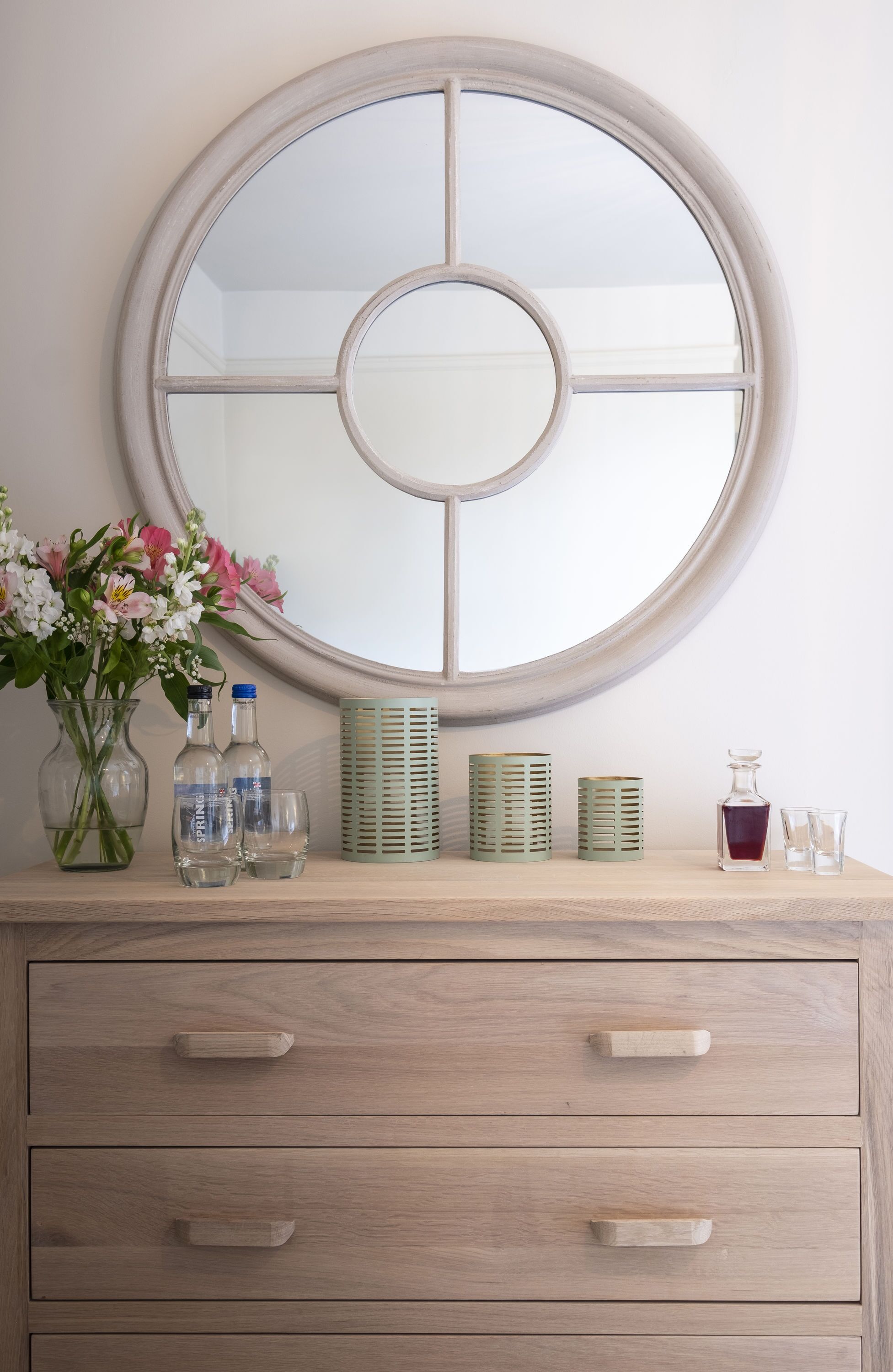 A wooden dresser with a round mirror above it, a vase with flowers, and several glass bottles and containers on the dresser.