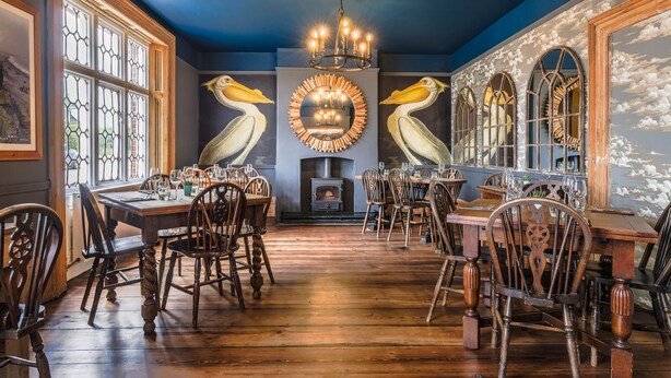 A dining area with wooden tables and chairs, a chandelier, and wall murals of pelicans. - The Ship at Dunwich (Saxmundham)
