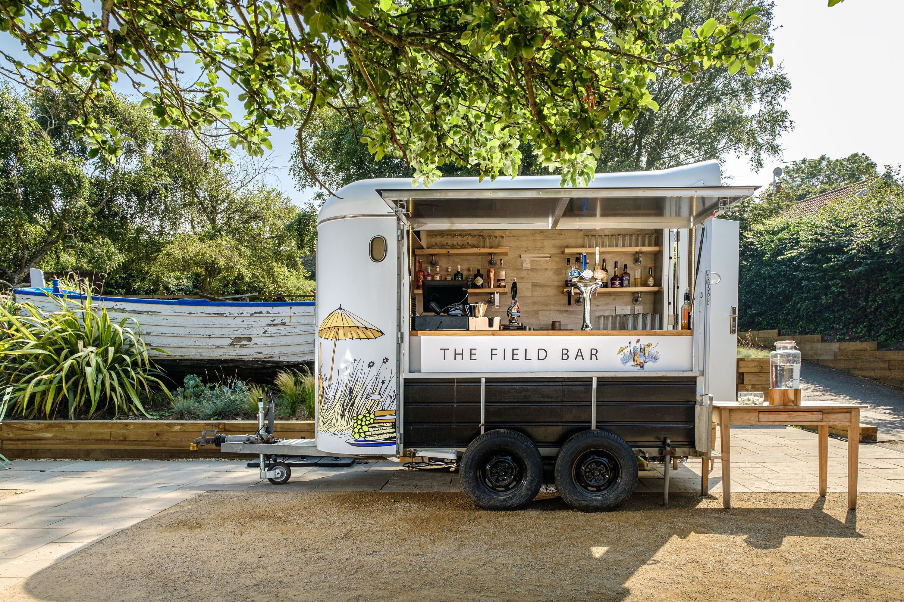 A mobile bar named "The Field Bar" with a trailer, a wooden table, and a garden setting.