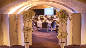 Un salón de banquetes interior con mesas dispuestas para un evento formal, una pantalla grande al frente y plantas en macetas en la entrada.