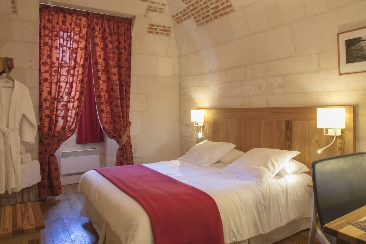 Une chambre avec un lit en bois, une couverture rouge, une table de chevet en bois, une chaise, un bureau en bois, un rideau rouge et une robe blanche suspendue à un crochet.